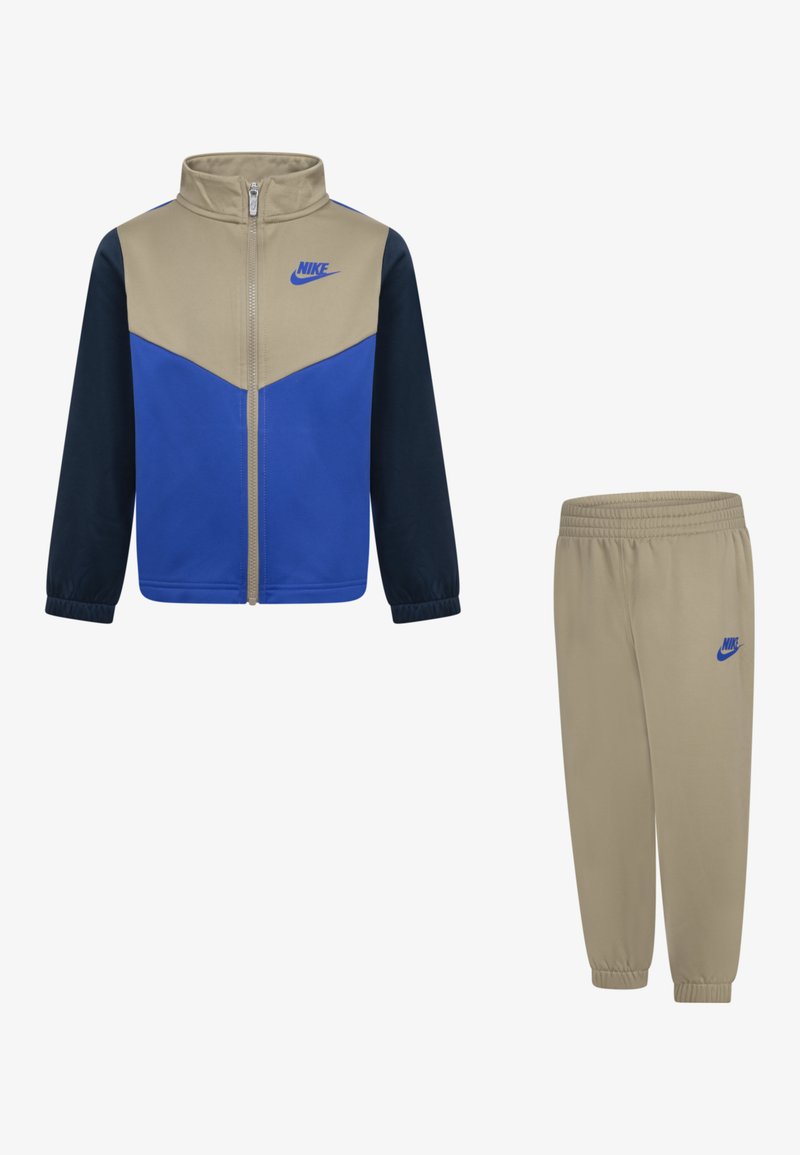 Nike trenerka, ki vključuje jakno z zadrgo v bež, modrih in mornarsko modrih barvah, v kombinaciji z bež hlačami z elastičnim pasom.