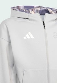 Haut de hoodie zippé gris clair avec un tissu texturé, présentant une doublure violette à motifs et des accents de logo réfléchissants sur la poitrine.