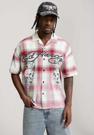 Młody mężczyzna z warkoczami, ubrany w czerwoną i białą kratowaną koszulę Ed Hardy z motywami czaszek i róż, dżinsy i czarną czapkę z haftem czaszki.