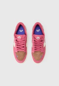 Nike SB sportcipő, rózsaszín velúr felsőrész, barna velúr orr rész perforálással, fehér középtalpak, és kék márkajelzés a talpbetéten és az oldalon.