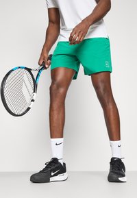 Grüne Tennishorts aus leichtem Material, kombiniert mit einem weißen Shirt, schwarzen Tennisschuhen und weißen Socken. Hält einen Tennisschläger.