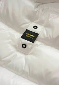 Tessuto trapuntato bianco con un'etichetta a pressione che presenta un'etichetta nera con la scritta "Blauer U.S.A." in testo giallo.