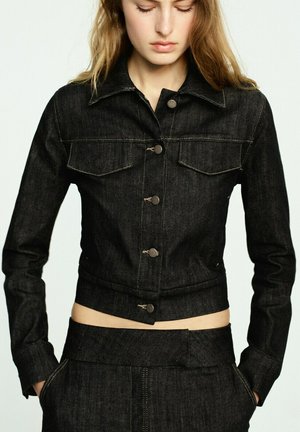 Woman wearing a fitted black denim jacket with button closure and matching high-waisted pants, hands in pockets, looking downward.