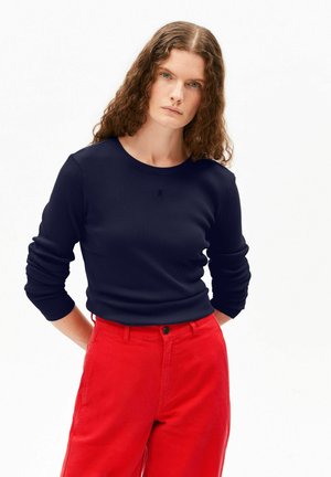 Femme aux longs cheveux bouclés portant un haut bleu marine à manches longues ajusté et un pantalon rouge taille haute, debout les mains derrière le dos.