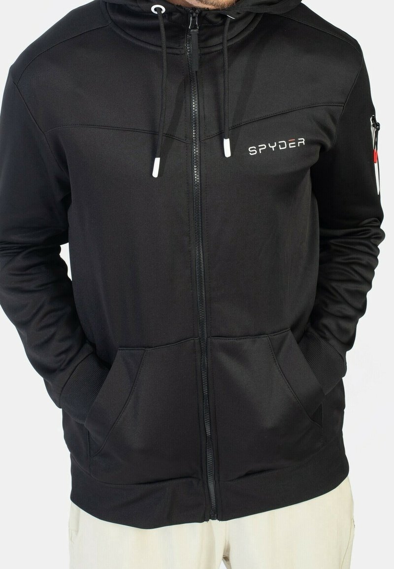 Homme portant un sweat à capuche zippé noir Spyder avec une poche avant et des cordons, mains dans les poches et pantalon beige.