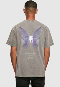 Mand iført falmet grå t-shirt med store lilla sommerfuglevinger og hvide stiliserede bogstaver på ryggen, stående mod en ensfarvet baggrund.