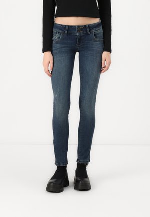 Slim fit donkerblauwe spijkerbroek gedragen met een zwart cropped langarmshirt en dikke zwarte platformschoenen op een witte achtergrond.