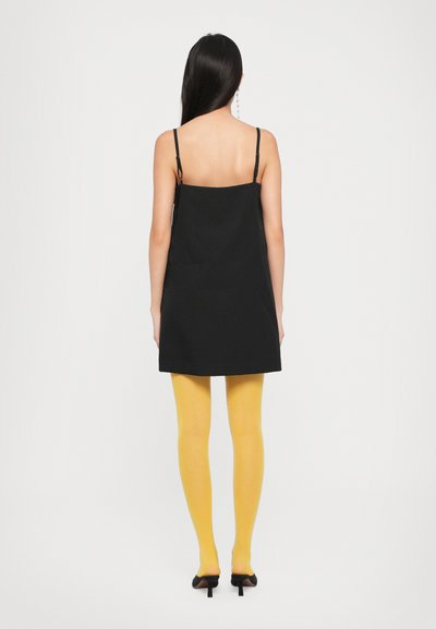 Fekete, ujjatlan ruha egyenes sziluettel, élénk sárga harisnyával párosítva. Átlátszó pántok és fekete magassarkú cipők is láthatók.