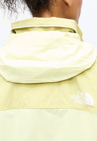 Γυναίκα που φορά ένα ανοιχτό κίτρινο μπουφάν North Face με κουκούλα διπλωμένη προς τα κάτω, δείχνοντας την πλάτη και το λογότυπο στο επάνω δεξί μέρος.