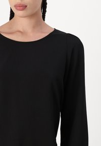 Haut noir à manches longues avec un col rond, fabriqué à partir d'un tissu lisse, présentant une texture subtile et un léger bombé aux coutures des épaules.