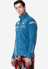 Giacca in pile blu con colletto alto, zip full e tasche con zip. Presenta loghi bianchi sul petto e sulla manica. Abbinata a pantaloni a fantasia.