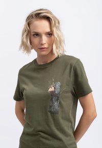 Olijfgroene T-shirt met een afbeelding van een boomstam met een vogelhuisje, bruine schorsstructuur en takken die naar buiten uitsteken. Korte mouwen.
