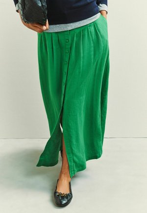 Person trägt einen grünen Midi-Rock mit Knopfleiste, schwarze Ballerinas mit goldenen Details, marineblaue und graue, geschichtete Oberteile und hält eine schwarze, geflochtene Handtasche.
