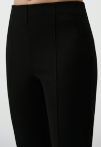 Zwarte taillerende broek met een zachte textuur, gestructureerde pasvorm en verticale naden. Zijzakken zijn in het ontwerp geïntegreerd.