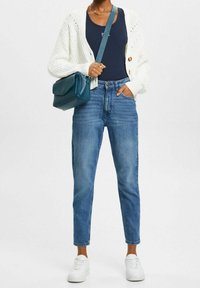 Vit stickad cardigan, marinblå linne, blå högmidjade jeans och vita sneakers med en teal färgad axelväska.