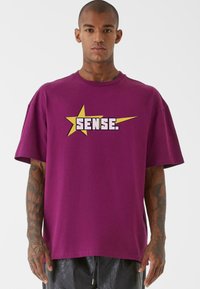 Fialové bavlněné tričko s velkou žlutou hvězdou a textem "SENSE." v bílé barvě, s krátkými rukávy a volným střihem.