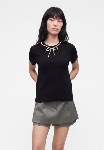 Femme aux longs cheveux noirs porte un haut noir à manches courtes avec un collier en perles en forme de nœud et un short gris texturé sur un fond uni.