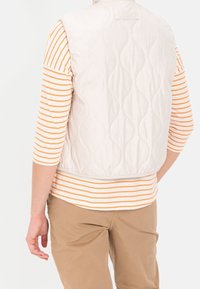 Vatteret hvid vest med en afrundet kant, båret over en langærmet stribet skjorte i hvid og orange. Beige bukser ses nedenfor.