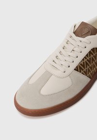 Bežiniai sportiniai bateliai su lygaus odos ir tekstūruotos zomšos deriniu, papuošti raštuota šonine panelė ir guma padanga.