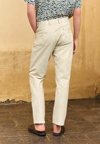 Pantalon en coton beige clair à coupe droite, avec deux poches au dos et des coutures visibles. Porté avec une chemise à motifs.