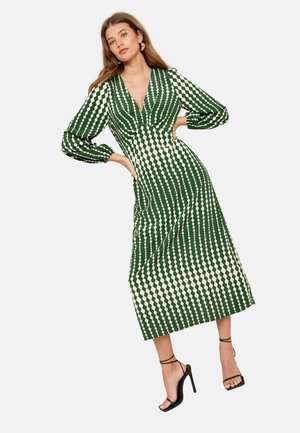 Ein grün- und cremefarbig gemustertes Kleid mit tiefem V-Ausschnitt, langen Puffärmeln und einer taillierten Silhouette. Kombiniert mit schwarzen, hochhackigen Sandalen.