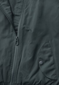 Giacca leggera di un verde scuro, con zip a lunghezza totale, dettaglio con bottone a pressione e tasca tonda dal tessuto morbido.