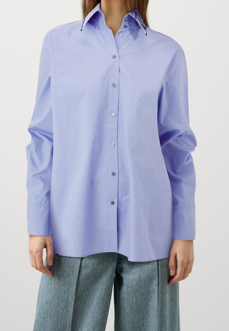 Camicia button-up lavanda con colletto a punta, maniche lunghe e bottoni argentati. Presenta una vestibilità rilassata e un tessuto liscio, leggermente lucido.
