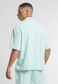 Camisa de manga corta con cuello en verde menta claro, con un patrón geométrico texturizado. Presenta un corte relajado y aberturas en los costados del dobladillo.