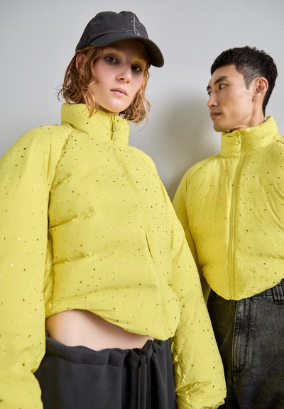 Yellow puffer jacket with a high collar, featuring a quilted design and scattered reflective accents. Styled with a black cap and grey pants.