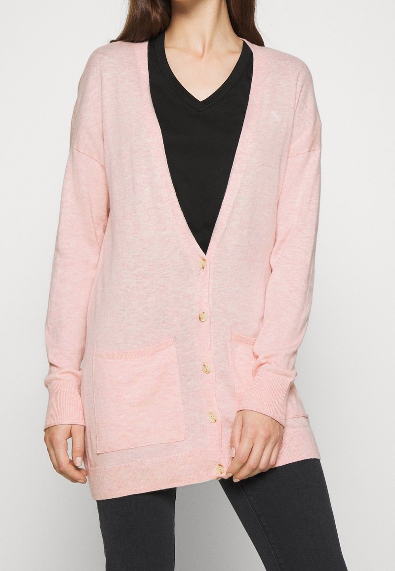 Světle růžový cardigan z měkkého materiálu s výstřihem do V, šesti knoflíky a dvěma předními kapsami. Lemované manžety a spodní okraj se žebrováním.