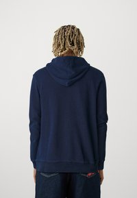 Marineblauwe hoodie met een trekkoord in de capuchon, ribbels bij de manchetten en onderkant. Gemaakt van een gestructureerde stof, met een subtiel logo aan de zijkant.