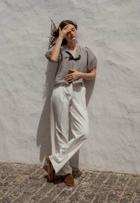 Blusa a righe a maniche corte, bianca e nera, con scollo tondo. Pantaloni bianchi a gamba larga, scarpe in suede marrone, contro un muro bianco testurizzato.