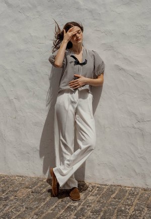 Blusa de manga corta a rayas, en blanco y negro, con escote redondo. Pantalones anchos blancos, zapatos de ante marrón, contra una pared texturizada blanca.
