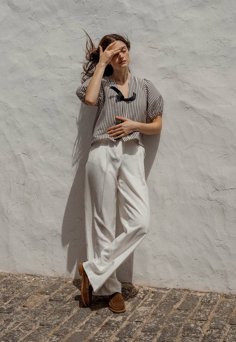 Blusa a righe a maniche corte, bianca e nera, con scollo tondo. Pantaloni bianchi a gamba larga, scarpe in suede marrone, contro un muro bianco testurizzato.