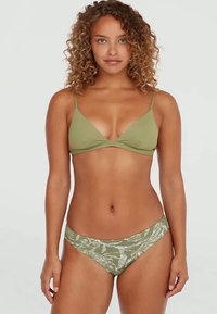 Ženska s kodrastimi lasmi, oblečena v zeleni bikini top in vzorčaste zelene bikini hlačke, stoji ob enobarvni svetli podlagi.