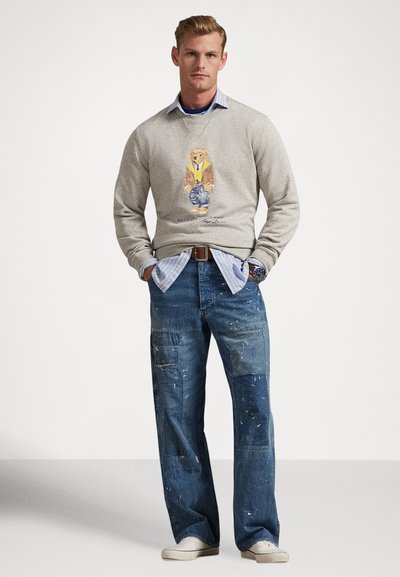 Γκρι φούτερ με γραφικό αρκούδας, φορεμένο πάνω από πουκάμισο με μπλε ρίγες. Συνδυασμένο με άνετο μπλε τζιν με πιτσιλιές από μπογιά και πλαϊνές τσέπες.