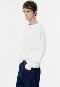 Weißer gerippter Pullover mit rundem Ausschnitt, lässiger Passform und langen Ärmeln, kombiniert mit dunkelblauen Jeans. Minimalistisches Design und Textur.