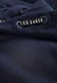 Navy sininen kangas, jossa on sileä rakenne, ja siinä on etiketti, jossa lukee "TED BAKER" valkoisella mustalla taustalla. Yksityiskohtina on kirjailtua koristeellisuutta.