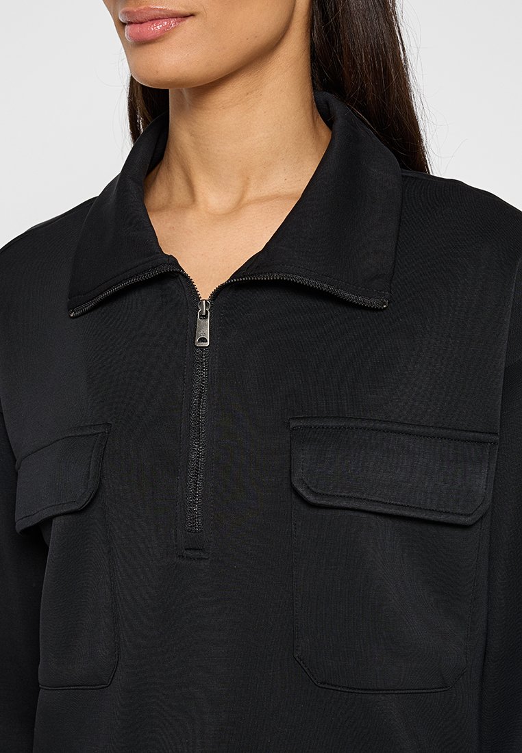 Schwarze Zip-Jacke mit umklappbarem Kragen, zwei Brusttaschen und einem strukturierten Stoff. Funktionales Design, geeignet für Freizeitkleidung.