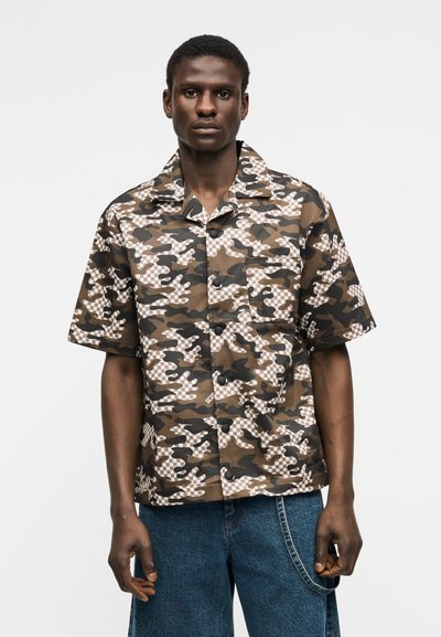 Νέος άνδρας που φοράει καφέ και μαύρο κοντομάνικο πουκάμισο παραλλαγής με καρό σχέδιο, συνδυασμένο με μπλε τζιν τζιν.