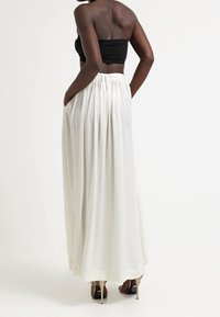 Bílá maxi sukně s nařaseným pasem, bočními kapsami a hladkou texturou; nošena s černým topem bez ramínek a sandály na podpatku.