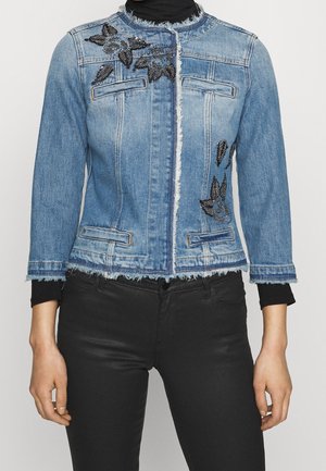 Mujer con chaqueta vaquera azul claro con bordado de cuentas florales negras, bordes deshilachados y pantalones negros.