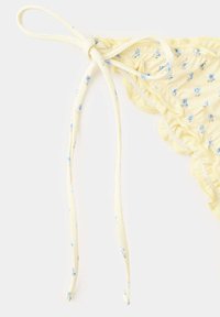 Bas de bikini jaune avec petites fleurs bleues et cordon latéral noué sur fond blanc.