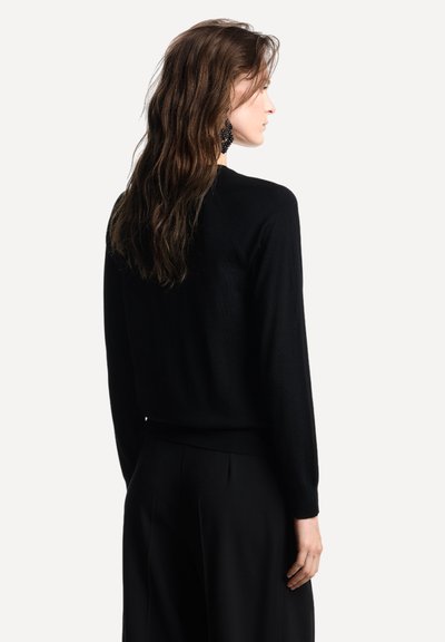 Maglione nero lavorato a maglia con maniche lunghe, scollo rotondo e polsini a coste. Tessuto morbido con silhouette aderente. Indossato con pantaloni neri a gamba larga.