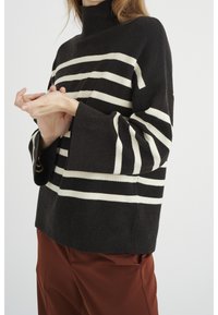 Suéter negro de cuello alto con rayas horizontales blancas, ajuste relajado y mangas anchas. Combinado con pantalones marrones. Textura suave.