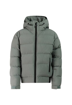 Jonge persoon glimlacht, draagt een groene winterjas, broek, muts, beige handschoenen, witte zonnebril en zwarte sneeuwlaarzen op een effen achtergrond.