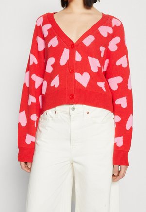 Femme portant un cardigan rouge boutonné avec des motifs de cœurs roses et un pantalon blanc taille haute sur un fond uni.