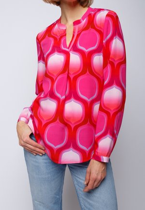 Femme portant un chemisier à manches longues avec un motif géométrique rose vif, rouge et blanc, et un jean bleu clair.