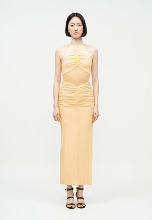 Femme aux cheveux noirs coiffés en carré, portant une robe halter pêche froncée sans manches et des talons noirs à brides, se tenant devant un fond blanc.