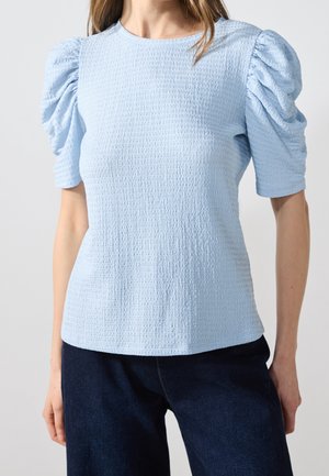 Femme portant un haut texturé bleu clair à manches courtes avec des épaules bouffantes et un pantalon bleu foncé, debout devant un fond uni.